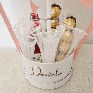 May include: A white gift box with the name "Daniela" written in gold lettering. The box is filled with a bottle of red wine, white tulle, and gold-wrapped chocolates.