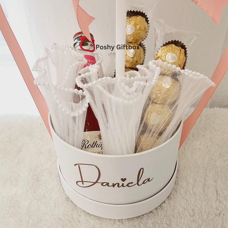 May include: A white gift box with the name "Daniela" written in gold lettering. The box is filled with a bottle of red wine, white tulle, and gold-wrapped chocolates.