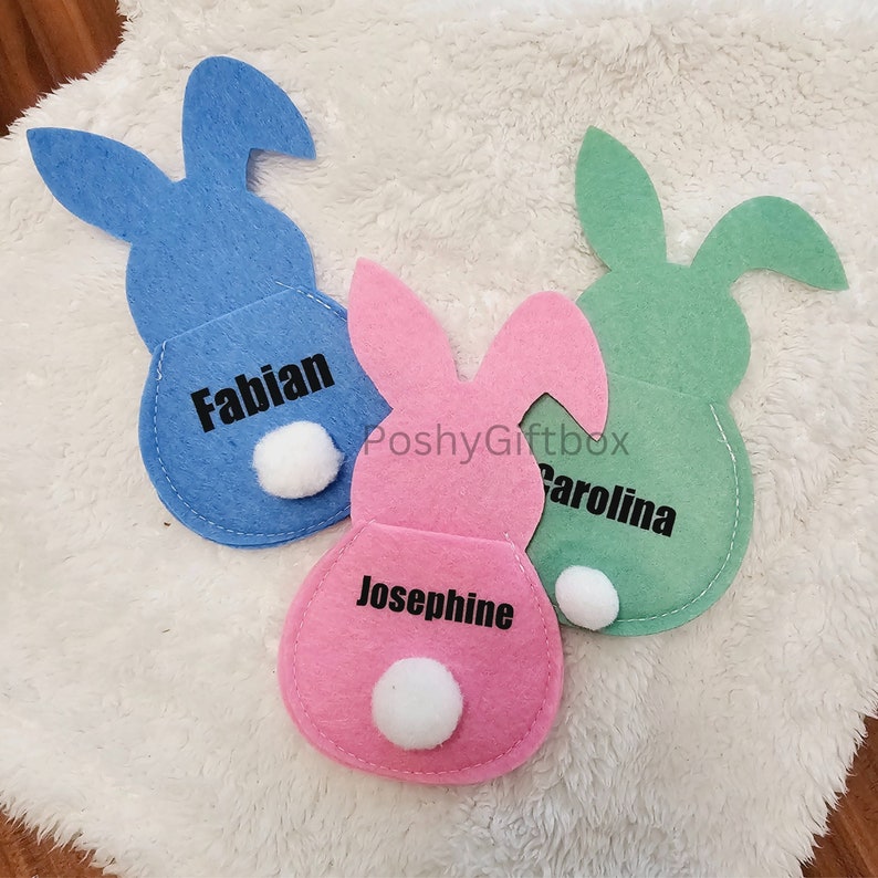 May include: Three felt bunny-shaped pouches with white pom-poms. The pouches are blue, pink, and green. The pouches are personalized with the names "Fabian", "Josephine", and "Carolina".