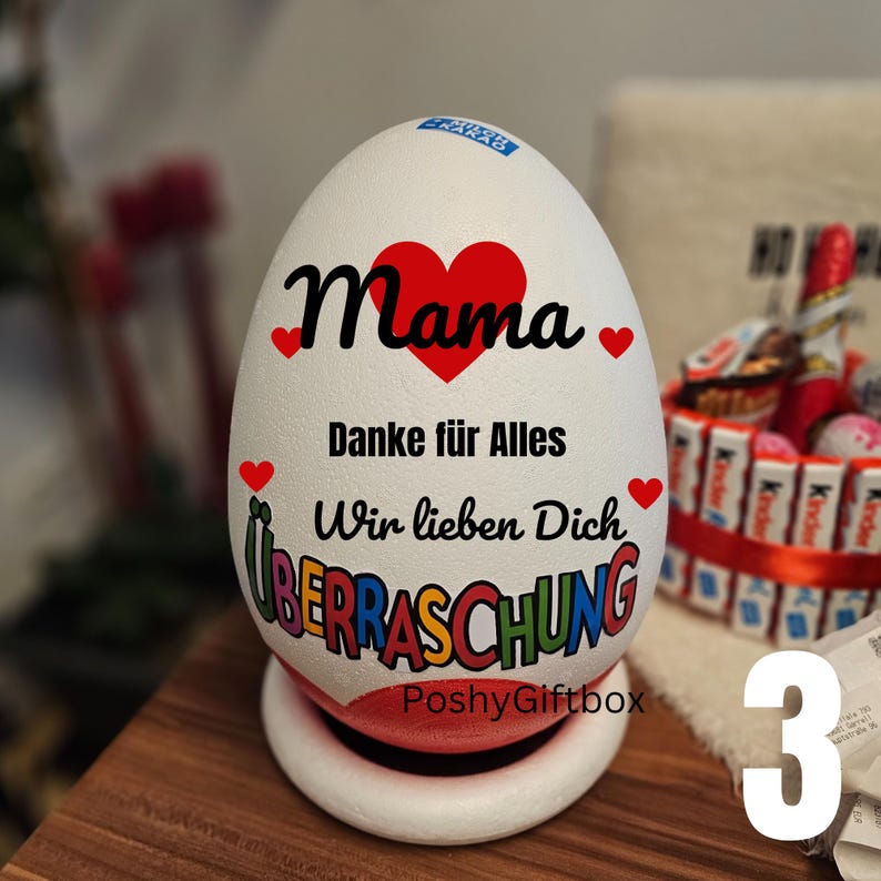 May include: A white egg-shaped gift box with the word "Mama" and a red heart. The text "Danke f&uuml;r Alles" and "Wir lieben Dich" are also visible. The word "&Uuml;BERRASCHUNG" is written in colorful letters. The base is white and red.