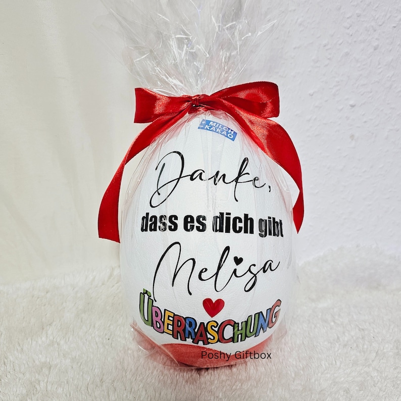 May include: A white egg-shaped gift box with a red ribbon and a red heart. The box is decorated with the text "Danke, dass es dich gibt Melisa &Uuml;berraschung" and the words "Poshy Giftbox".