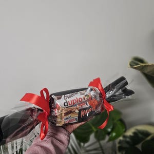 May include: A cylindrical gift package wrapped in clear cellophane with red ribbon bows tied at both ends. The centre contains assorted chocolate bars including Kinder Bueno, Duplo, and Hanuta Minis, visible through the transparent plastic wrapping.
