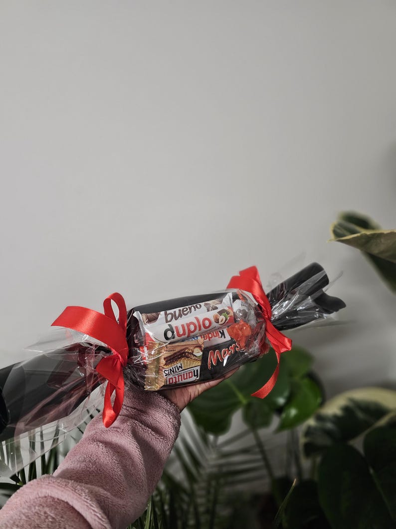May include: A cylindrical gift package wrapped in clear cellophane with red ribbon bows tied at both ends. The centre contains assorted chocolate bars including Kinder Bueno, Duplo, and Hanuta Minis, visible through the transparent plastic wrapping.