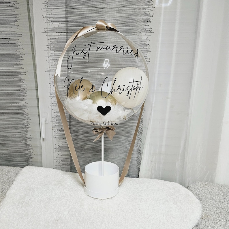 May include: A clear balloon hot air balloon with a white and gold balloon inside. The balloon is tied to a white box with a brown ribbon. The balloon has the text "Just married" and "Nile & Christoph" written on it. The balloon is on a white surface.