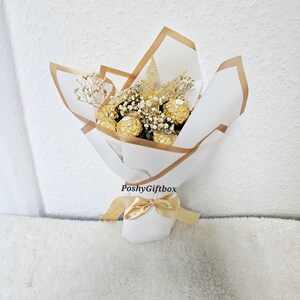 May include: A bouquet of gold-wrapped chocolate candies with dried white flowers and gold ribbon. The bouquet is wrapped in white paper with a gold border and the text "PoshyGiftbox" is printed on the paper.