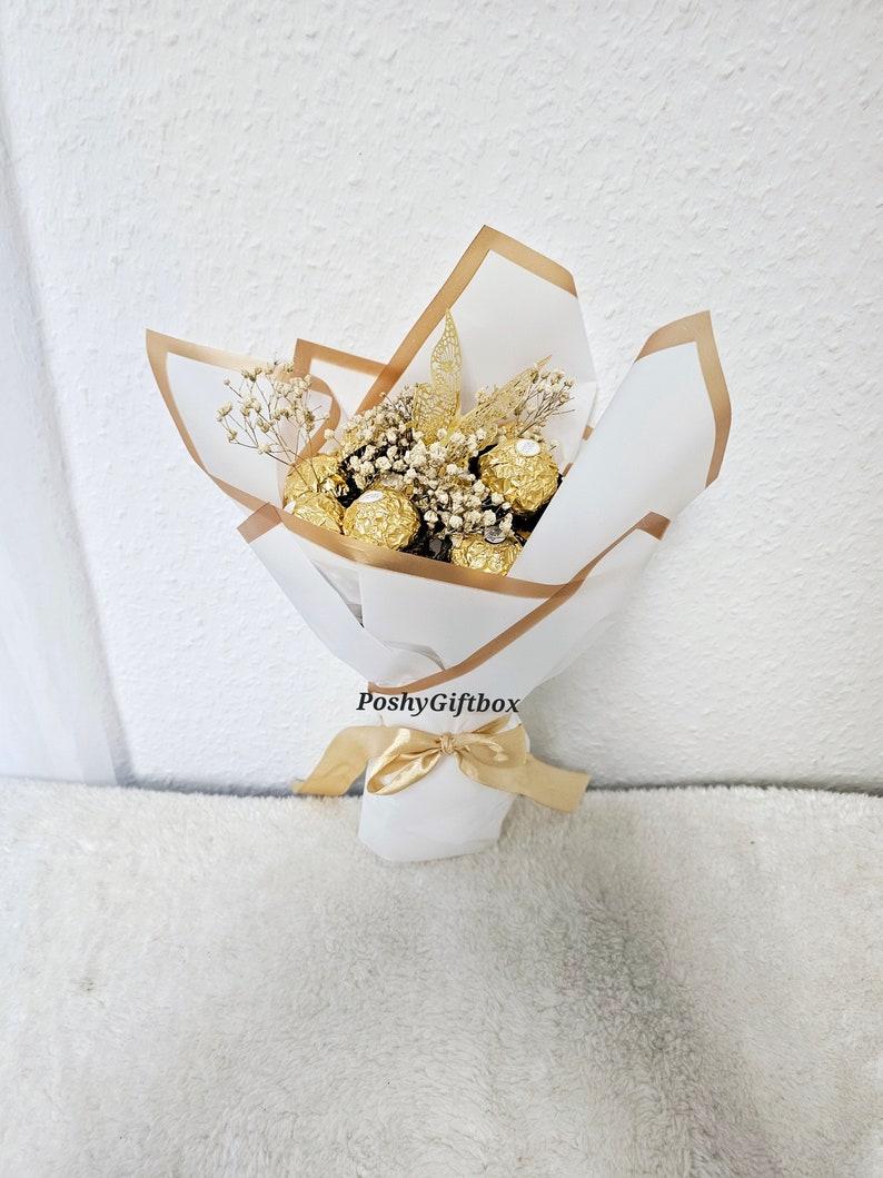 May include: A bouquet of gold-wrapped chocolate candies with dried white flowers and gold ribbon. The bouquet is wrapped in white paper with a gold border and the text "PoshyGiftbox" is printed on the paper.