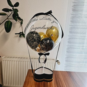 Op de afbeelding: Een doorzichtige ballon gevuld met kleinere zwarte en gouden ballonnen, voorzien van de tekst Alles Liebe zur Jugendweihe in zwarte letters. De ballon is bevestigd op een hartvormige zwarte doos met een witte stok en zwarte linten.