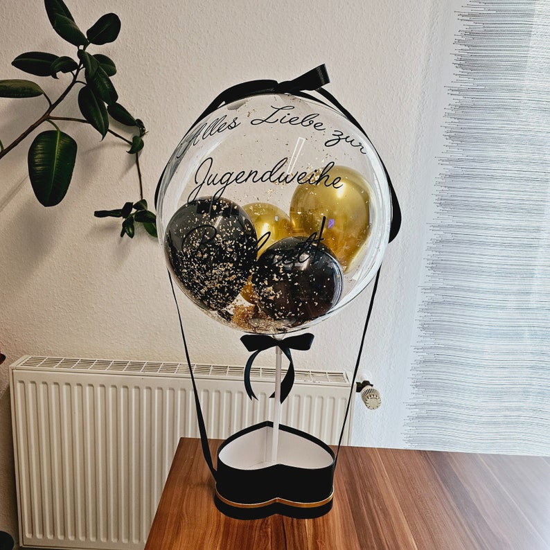 Op de afbeelding: Een doorzichtige ballon gevuld met kleinere zwarte en gouden ballonnen, voorzien van de tekst Alles Liebe zur Jugendweihe in zwarte letters. De ballon is bevestigd op een hartvormige zwarte doos met een witte stok en zwarte linten.