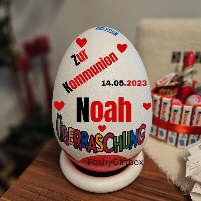 May include: A white egg-shaped gift box with red hearts and text. The text includes "Zur Kommunion", "14.05.2023", "Noah", and "&Uuml;BERRASCHUNG". The box sits on a white and red base, with the shop name "PoshyGiftbox" at the bottom.