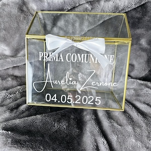 May include: A clear glass box with a gold trim and a white ribbon. The box has the text "Prima Comunione Aurelia Zerone 04.05.2025" printed on it.