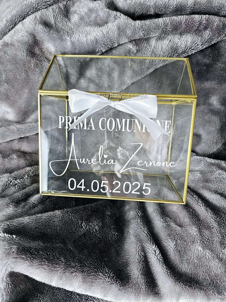 May include: A clear glass box with a gold trim and a white ribbon. The box has the text "Prima Comunione Aurelia Zerone 04.05.2025" printed on it.