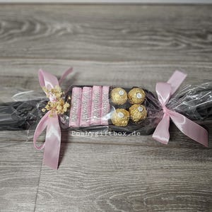 May include: A gift-wrapped confectionery assortment shaped like a cracker, featuring four pink Yogurette chocolate bars and four gold-wrapped Ferrero Rocher truffles. The package is wrapped in black paper, clear cellophane, and tied with light pink satin ribbons.