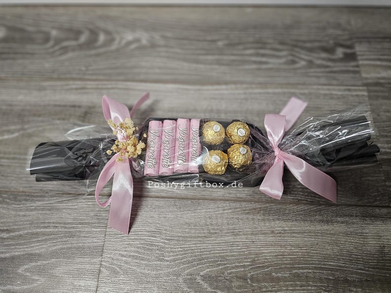 May include: A gift-wrapped confectionery assortment shaped like a cracker, featuring four pink Yogurette chocolate bars and four gold-wrapped Ferrero Rocher truffles. The package is wrapped in black paper, clear cellophane, and tied with light pink satin ribbons.