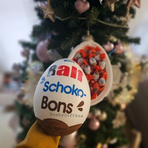 Peut inclure: Un &oelig;uf en chocolat blanc avec les mots "Rall Schoko-Bons PoshyGiftbox" imprim&eacute;s dessus. L'&oelig;uf est ouvert et rempli de petits bonbons au chocolat.