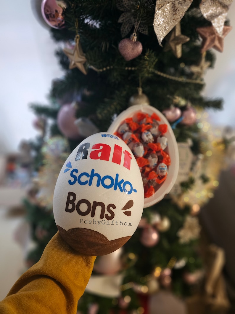 Peut inclure: Un &oelig;uf en chocolat blanc avec les mots "Rall Schoko-Bons PoshyGiftbox" imprim&eacute;s dessus. L'&oelig;uf est ouvert et rempli de petits bonbons au chocolat.