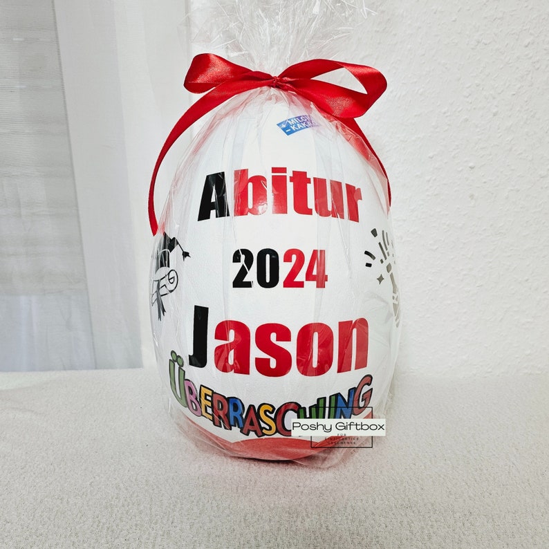 May include: A white and red surprise egg with the text "Abitur 2024 Jason" printed on it. The egg is wrapped in clear plastic and tied with a red ribbon.
