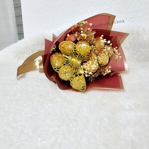 May include: A bouquet of chocolate candies wrapped in red and gold foil, with a gold butterfly decoration and dried white flowers. The bouquet is wrapped in red tissue paper with a gold ribbon and the text "PoshyGiftbox" printed on the paper.