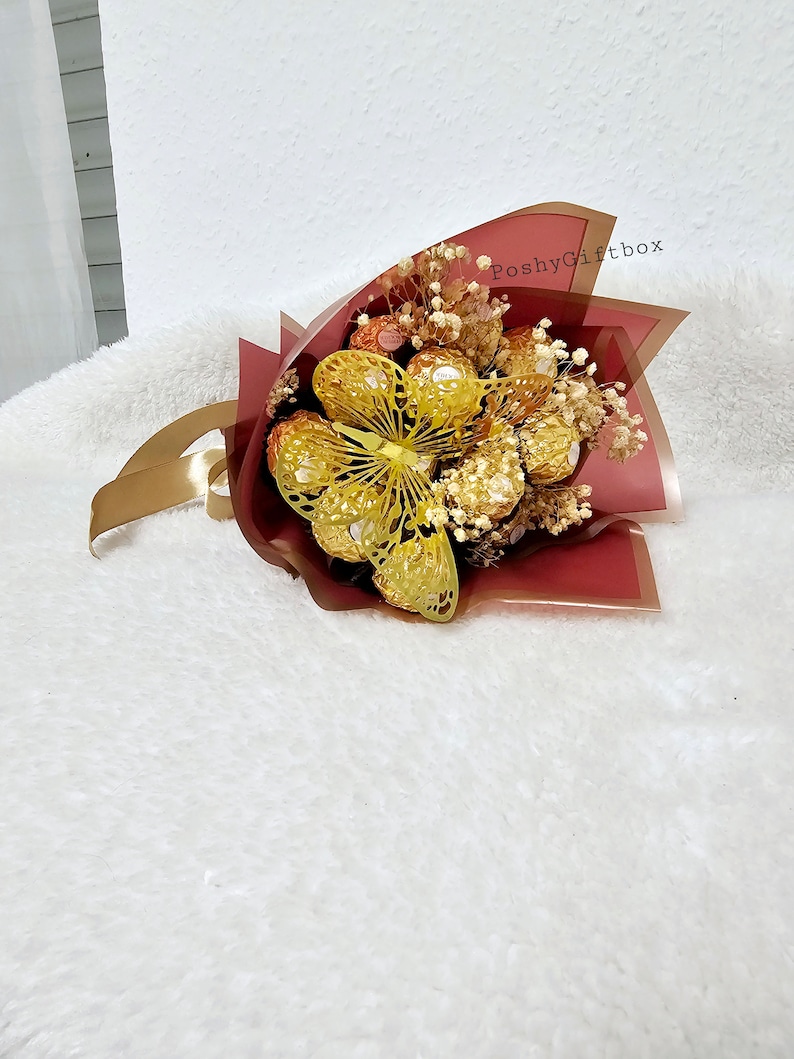 May include: A bouquet of chocolate candies wrapped in red and gold foil, with a gold butterfly decoration and dried white flowers. The bouquet is wrapped in red tissue paper with a gold ribbon and the text "PoshyGiftbox" printed on the paper.