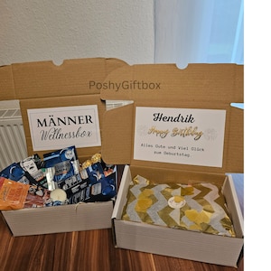 May include: Two gift boxes, one with a brown cardboard exterior and the other with a white exterior. The brown box has a white label that says "M&auml;nner Wellnessbox" and the white box has a white label that says "Hendrik Happy Birthday Alles Gute und Viel Gl&uuml;ck zum Geburtstag."