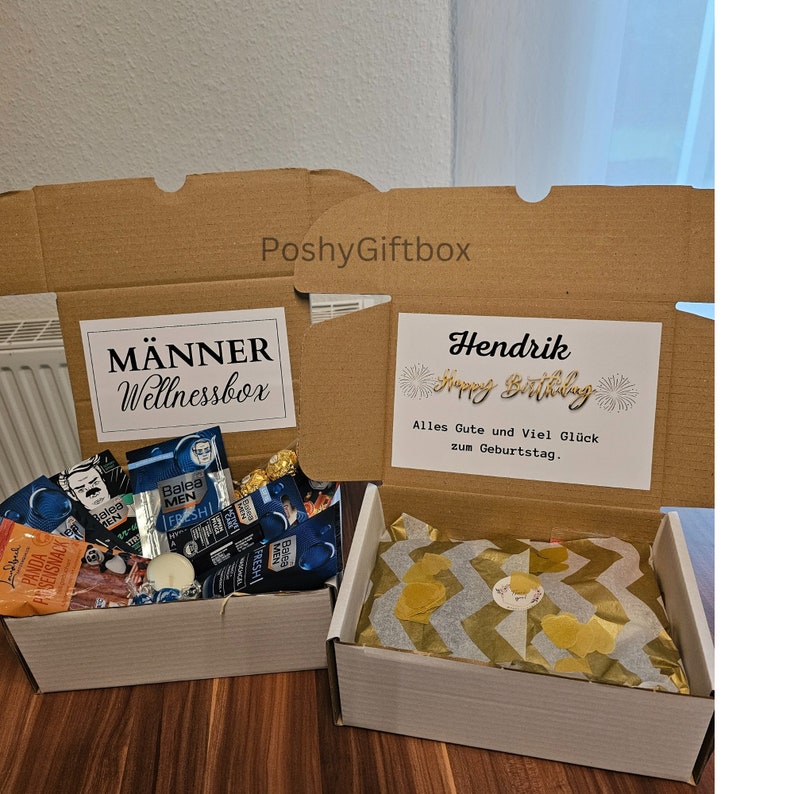 May include: Two gift boxes, one with a brown cardboard exterior and the other with a white exterior. The brown box has a white label that says "M&auml;nner Wellnessbox" and the white box has a white label that says "Hendrik Happy Birthday Alles Gute und Viel Gl&uuml;ck zum Geburtstag."