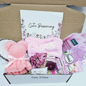 Peut inclure: Une bo&icirc;te cadeau rose remplie d'articles de soins personnels, dont un bandeau rose, un ours en peluche rose, une poudre de bain rose, une cr&egrave;me pour les mains rose, une bougie rose et une &eacute;ponge de bain rose. La bo&icirc;te est &eacute;tiquet&eacute;e "Poshy Giftbox" et porte une carte qui dit "Gute Besserung", ce qui signifie "Bon r&eacute;tablissement" en allemand.