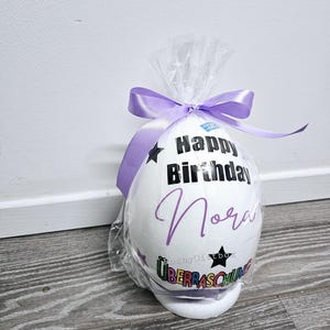 May include: A white foam egg with a purple ribbon, decorated with the text "Happy Birthday Nora" and a rainbow colored "Ueberraschung" sticker.