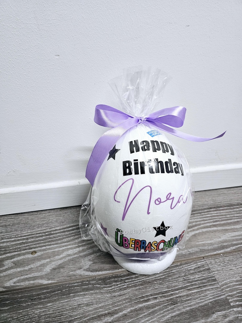May include: A white foam egg with a purple ribbon, decorated with the text "Happy Birthday Nora" and a rainbow colored "Ueberraschung" sticker.