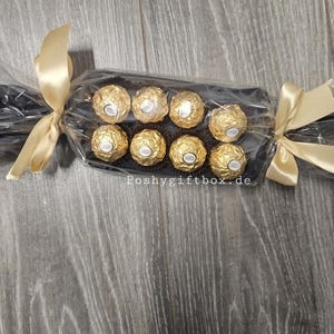 May include: A gift package shaped like a large sweet wrapper containing eight gold-foiled chocolates arranged in two rows. The clear cellophane wrapping is tied with gold satin ribbons at both ends. The text Poshygiftbox.de is visible.