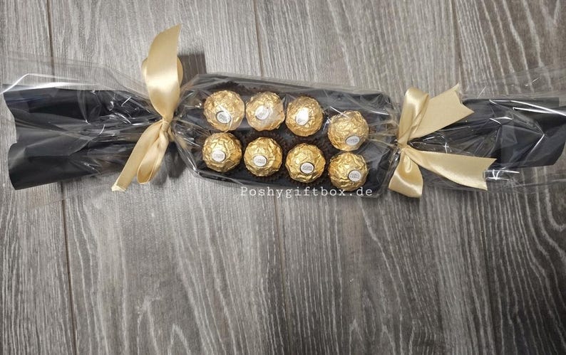 May include: A gift package shaped like a large sweet wrapper containing eight gold-foiled chocolates arranged in two rows. The clear cellophane wrapping is tied with gold satin ribbons at both ends. The text Poshygiftbox.de is visible.
