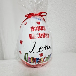 May include: White egg-shaped balloon with a red ribbon and the text "Happy Birthday Leni 18" in black. The balloon is wrapped in clear plastic and has a red heart on the side. The bottom of the balloon has the text "&Uuml;berraschung" in colorful letters and the text "PoshyGiftbox" in black.