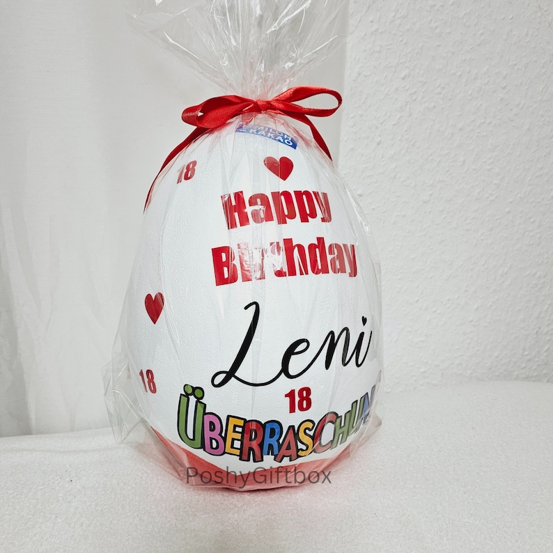May include: White egg-shaped balloon with a red ribbon and the text "Happy Birthday Leni 18" in black. The balloon is wrapped in clear plastic and has a red heart on the side. The bottom of the balloon has the text "&Uuml;berraschung" in colorful letters and the text "PoshyGiftbox" in black.