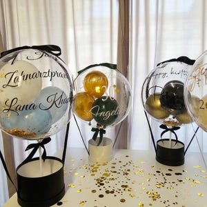 Op de afbeelding: Drie doorzichtige, bolvormige ballonnen gevuld met kleinere ballonnen in wit, blauw, goud, groen en zwart, rustend op ronde zwarte en witte dozen. De ballonnen zijn voorzien van zwarte en gouden cursieve tekst met namen en datums.
