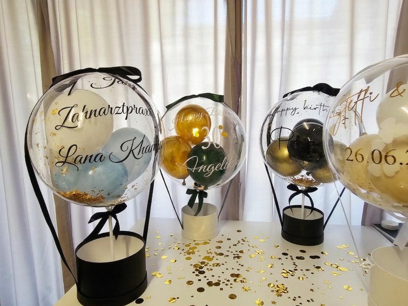 Op de afbeelding: Drie doorzichtige, bolvormige ballonnen gevuld met kleinere ballonnen in wit, blauw, goud, groen en zwart, rustend op ronde zwarte en witte dozen. De ballonnen zijn voorzien van zwarte en gouden cursieve tekst met namen en datums.