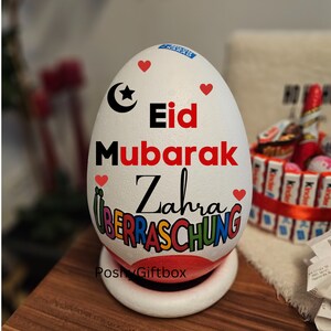 May include: White egg-shaped gift box with a red, white, and blue design. The box is decorated with the words "Eid Mubarak Zahra &Uuml;berraschung" and a crescent moon and star. The box is sitting on a white base.