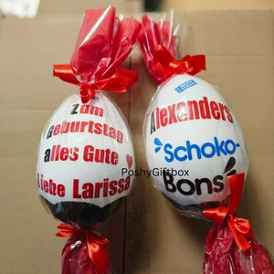Peut inclure: Deux &oelig;ufs en chocolat blanc envelopp&eacute;s de cellophane rouge avec des rubans rouges. Un &oelig;uf porte le texte "Zum Geburtstag alles Gute liebe Larissa" et un c&oelig;ur rouge. L'autre &oelig;uf porte le texte "Alexanders Schoko-Bons" avec un logo bleu et blanc.