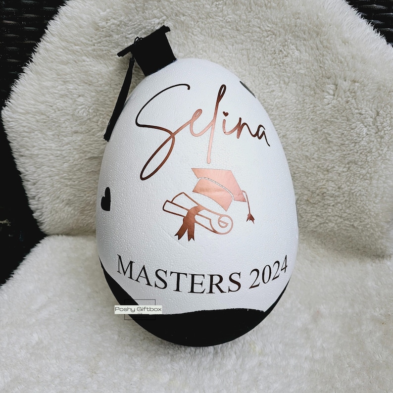 May include: White egg-shaped ornament with a black graduation cap on top. The ornament is decorated with a gold inscription that reads "Selina Masters 2024". There is a gold graduation cap and diploma design on the ornament.