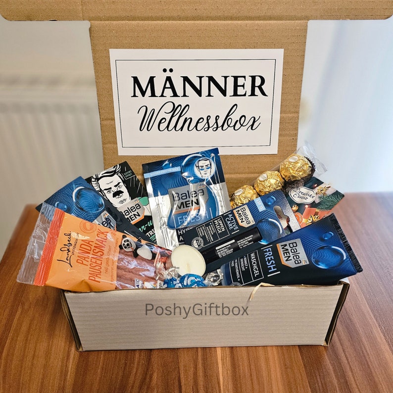 May include: A brown cardboard box with the text "M&Auml;NNER Wellnessbox" on the lid. The box is filled with various items, including a tube of face wash, a package of panda-shaped snacks, a candle, and a few chocolate candies.