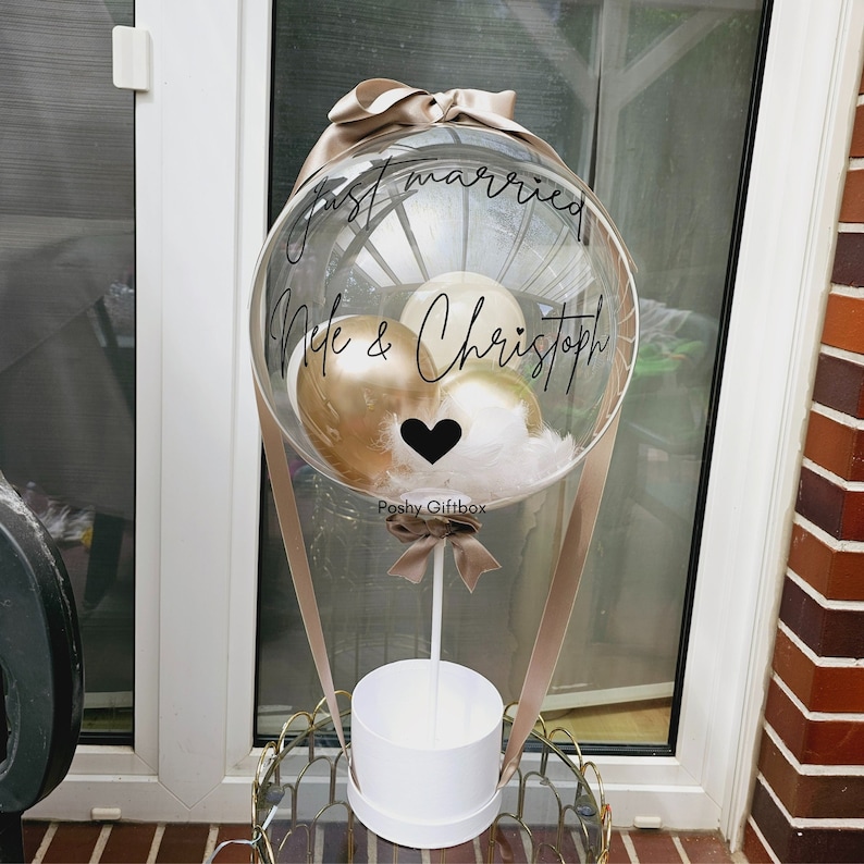 May include: A clear balloon with a white base and beige ribbon, filled with gold and white balloons and feathers. The balloon has the text "Just married" and "Nele & Christoph" written on it. The balloon is sitting on a white box with the text "Poshy Giftbox" written on it.