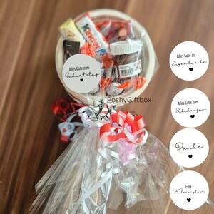 May include: A white bowl filled with various candies and chocolate bars, wrapped in clear cellophane and tied with red, white, and pink ribbons. The bowl is attached to a wooden stick and decorated with circular paper tags with text in German, including "Alles Gute zum Geburtstag" and "Danke".