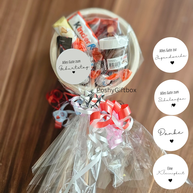 May include: A white bowl filled with various candies and chocolate bars, wrapped in clear cellophane and tied with red, white, and pink ribbons. The bowl is attached to a wooden stick and decorated with circular paper tags with text in German, including "Alles Gute zum Geburtstag" and "Danke".