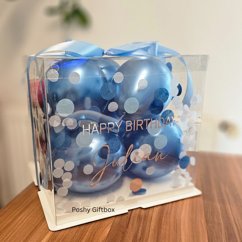 May include: A clear plastic box filled with blue balloons and confetti. The box has a white base and a blue ribbon. The text "Happy Birthday Julian" is printed on the box.