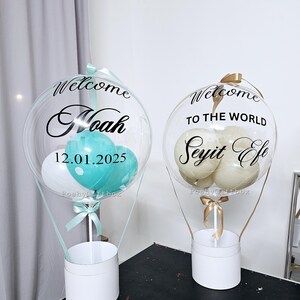 May include: Two clear balloon hot air balloon arrangements with white and colored balloons inside. The balloons are decorated with black text. One balloon says "Welcome Noah 12.01.2025" and the other says "Welcome to the world Seyit Efe".