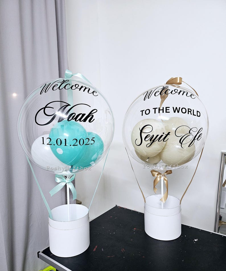 May include: Two clear balloon hot air balloon arrangements with white and colored balloons inside. The balloons are decorated with black text. One balloon says "Welcome Noah 12.01.2025" and the other says "Welcome to the world Seyit Efe".