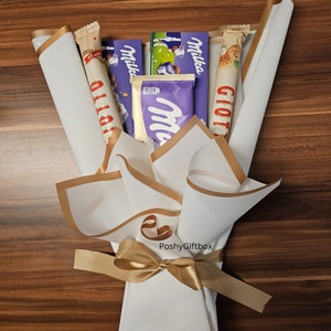 May include: A white and gold gift bouquet with a gold ribbon. The bouquet contains various chocolate bars, including Milka, Giotto, and a white chocolate bar with a purple wrapper.