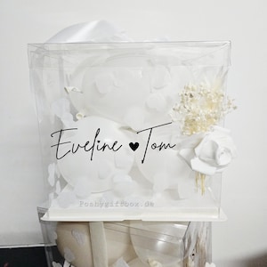 May include: A clear, square gift box filled with white balloons, paper cutouts, and floral accents. The box is adorned with the names "Eveline & Tom" in elegant script. The website "Poshygiftbox.de" is printed on the box.