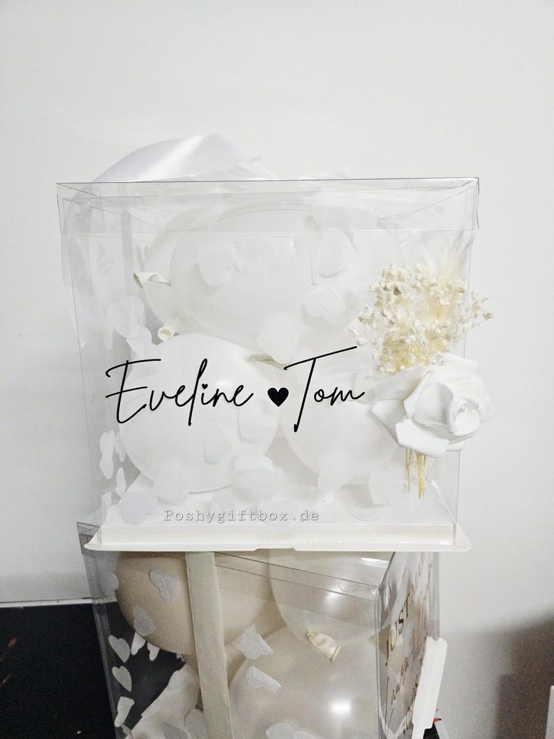 May include: A clear, square gift box filled with white balloons, paper cutouts, and floral accents. The box is adorned with the names "Eveline & Tom" in elegant script. The website "Poshygiftbox.de" is printed on the box.
