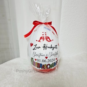 以下が含まれることがあります： 赤いリボンと「Zur Hochzeit Christiane & Christoph 01.06.2024 &Uuml;berraschung」というメッセージが印刷された白い風船。風船は赤いハートと2羽の赤い鳥で飾られています。