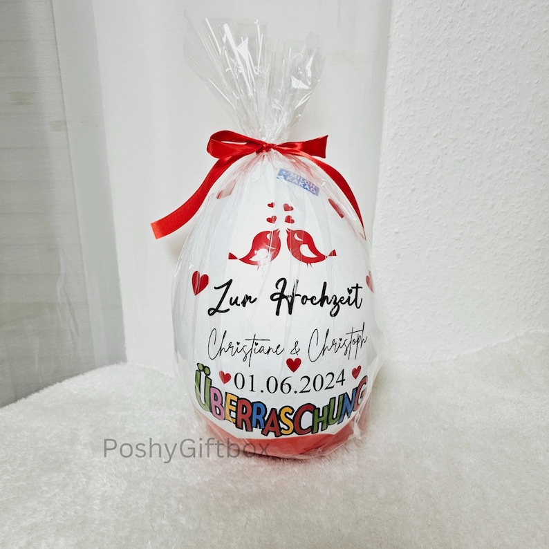 以下が含まれることがあります： 赤いリボンと「Zur Hochzeit Christiane & Christoph 01.06.2024 &Uuml;berraschung」というメッセージが印刷された白い風船。風船は赤いハートと2羽の赤い鳥で飾られています。