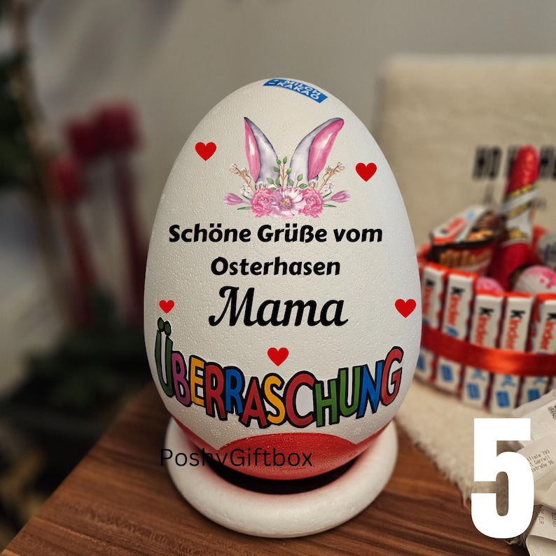 May include: A white egg-shaped decorative item with text in German, featuring bunny ears, hearts, and the word "Mama." The egg sits on a white base with a red bottom. The word "&Uuml;BERRASCHUNG" is also visible.