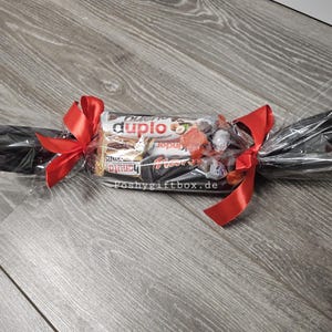 May include: A clear cellophane gift wrapper shaped like a large sweet, containing assorted chocolate bars including Duplo and Kinder products. The ends are twisted and secured with bright red satin ribbons. The text poshygiftbox.de is visible.